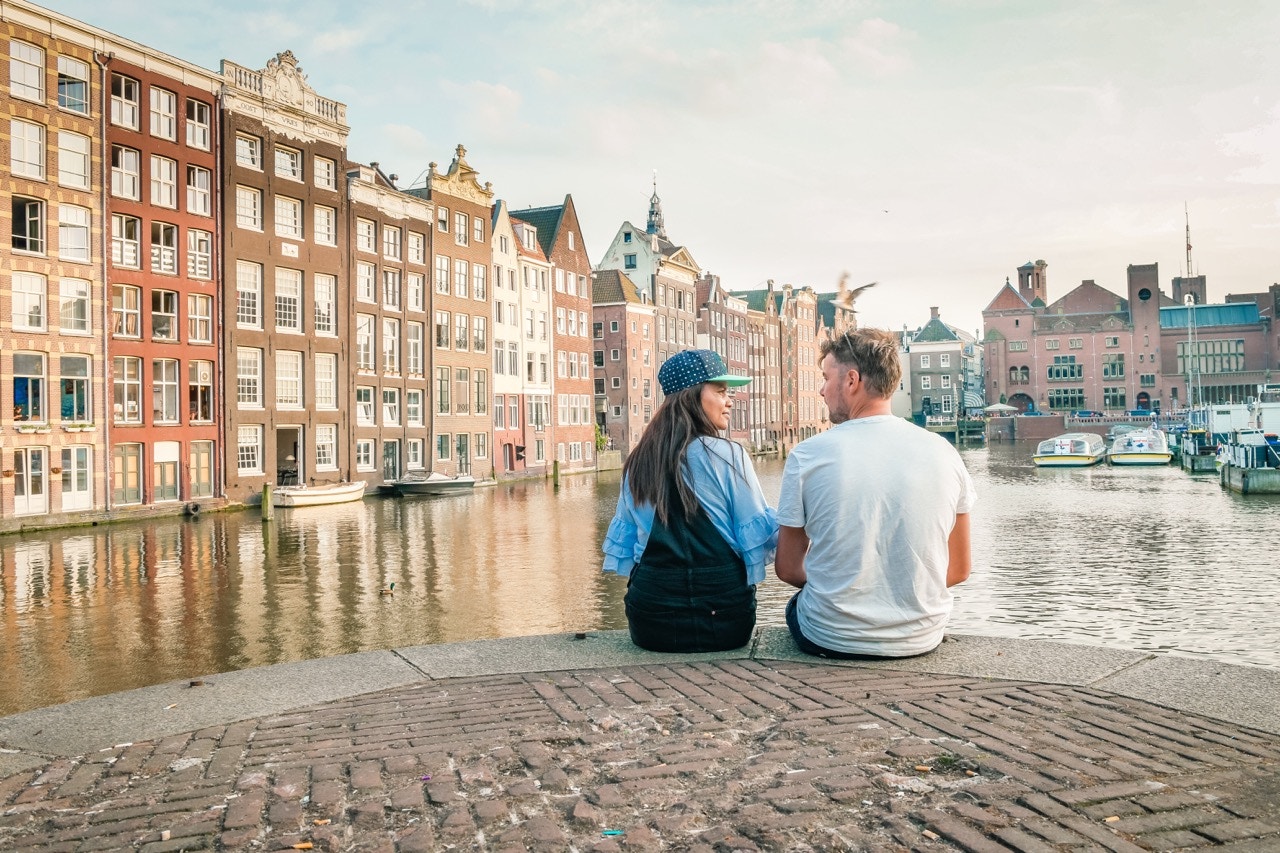 Couple sitting by Amsterdam's Damrak canal with historic buildings in view.