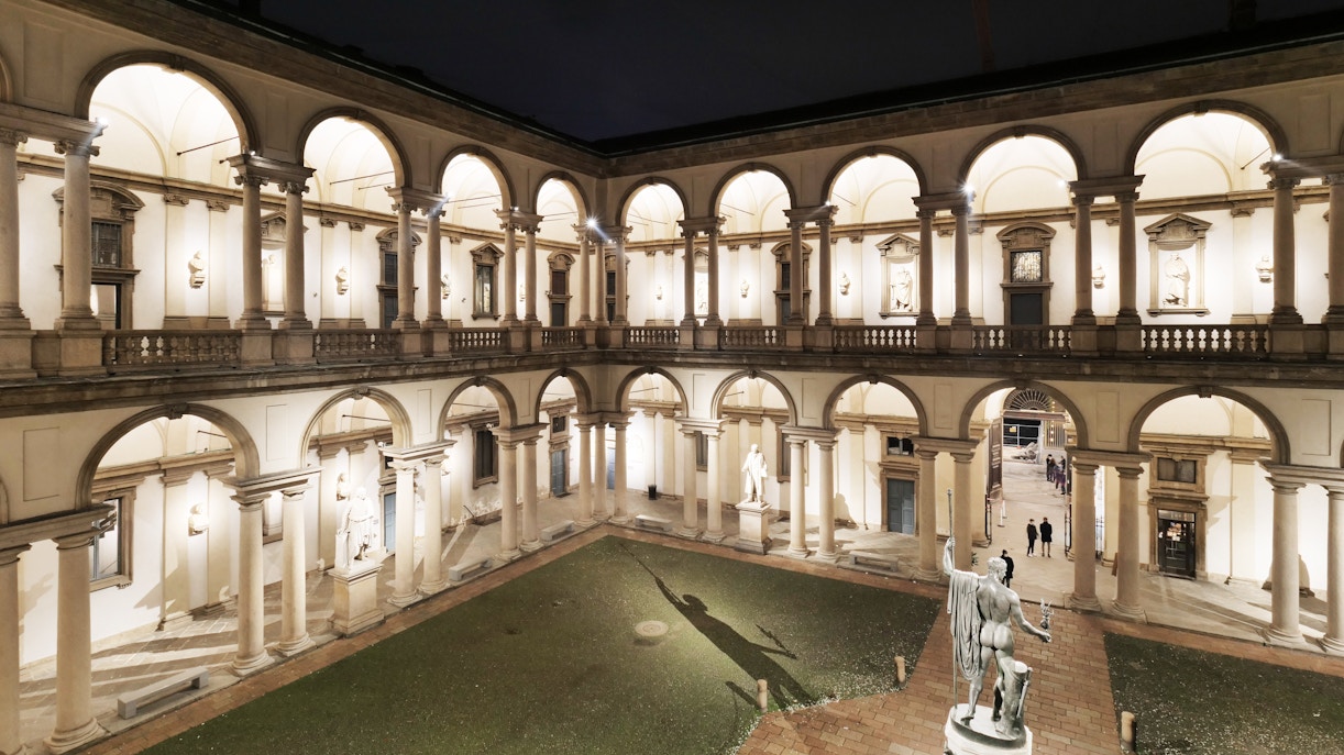 Pinacoteca di Brera courtyard at night, Milan, with illuminated arches and statue.