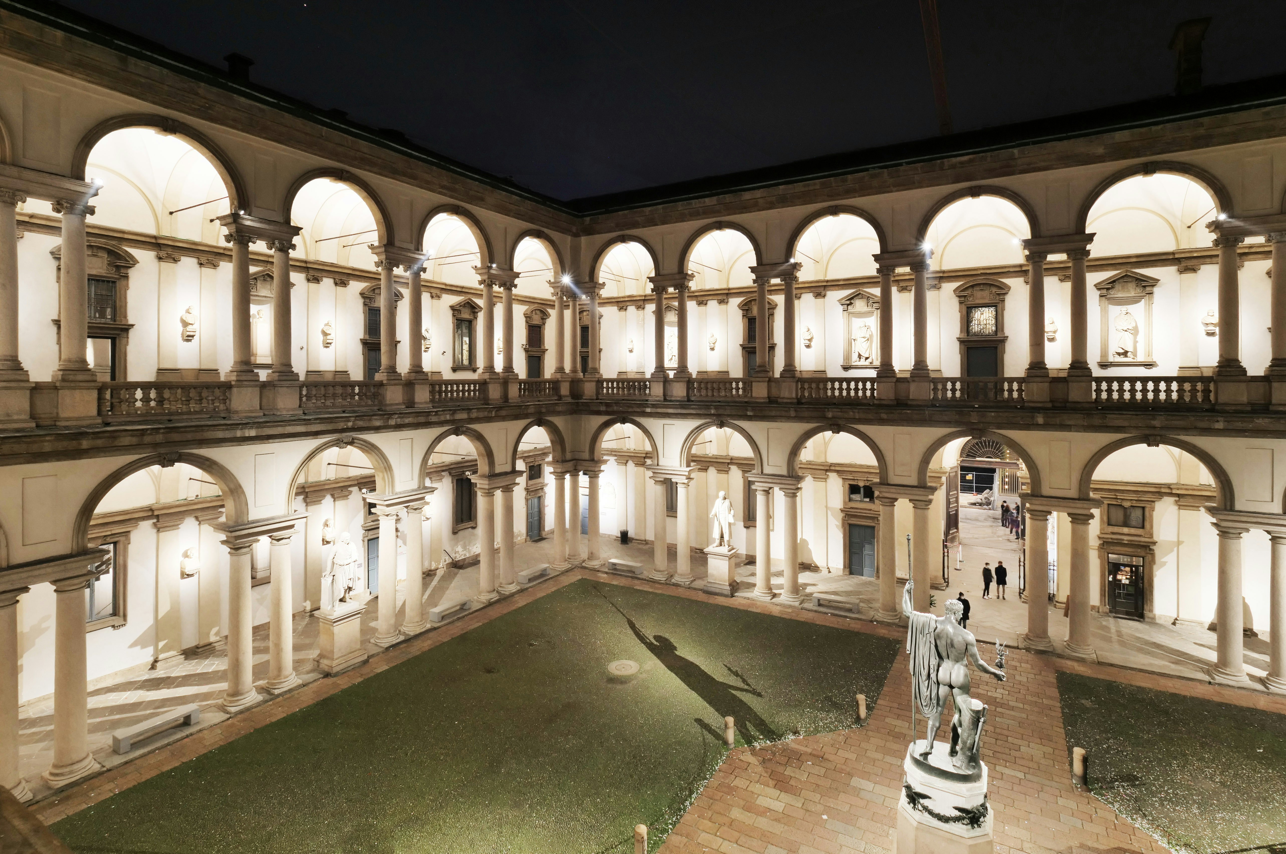 Pinacoteca di Brera courtyard at night, Milan, with illuminated arches and statue.