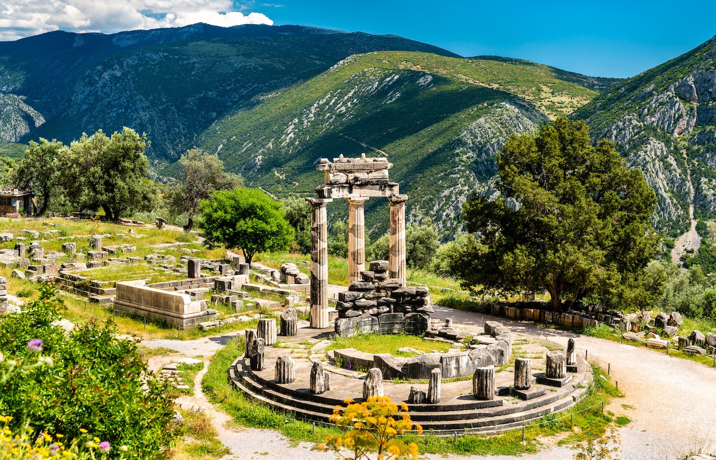 Visitors at the Temple of Apollo in Delphi, Greece, surrounded by ancient ruins and mountains.