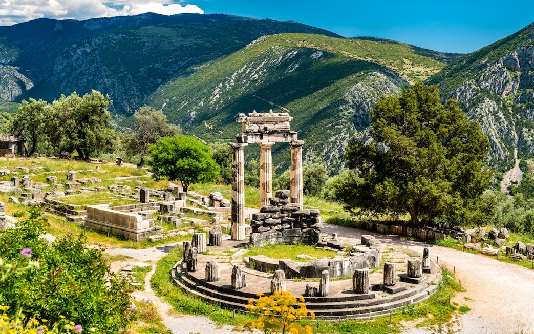 Visitors at the Temple of Apollo in Delphi, Greece, surrounded by ancient ruins and mountains.