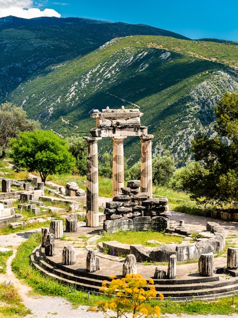 Visitors at the Temple of Apollo in Delphi, Greece, surrounded by ancient ruins and mountains.