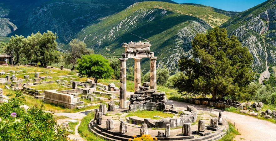 Visitors at the Temple of Apollo in Delphi, Greece, surrounded by ancient ruins and mountains.