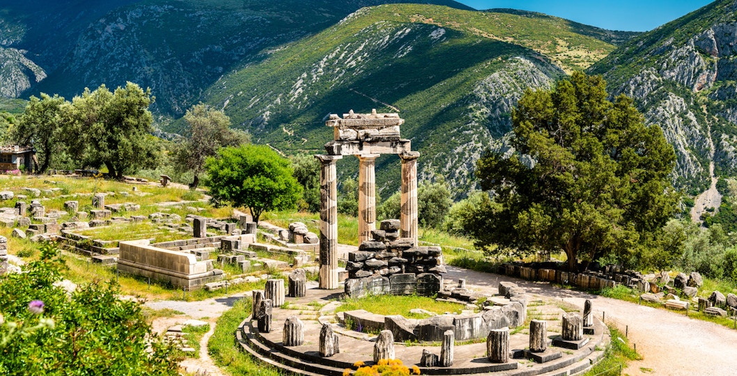 Visitors at the Temple of Apollo in Delphi, Greece, surrounded by ancient ruins and mountains.