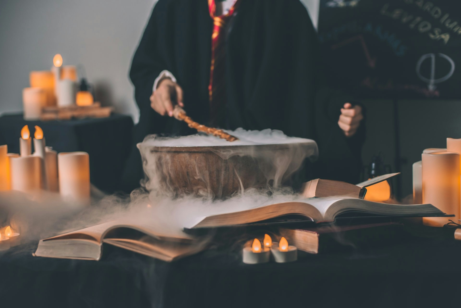 Person in wizard robe stirring cauldron with wand, surrounded by candles and open books.