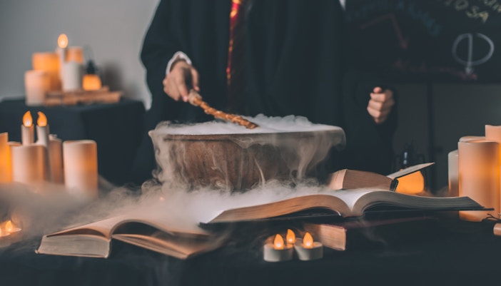 Person in wizard robe stirring cauldron with wand, surrounded by candles and open books.
