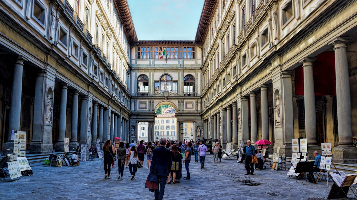 Os visitantes se aproximam da grande entrada ingresso Galeria Uffizi e caminham por um elegante pátio revestido de colunas e arcos clássicos, mostrando o rico patrimônio de Florença