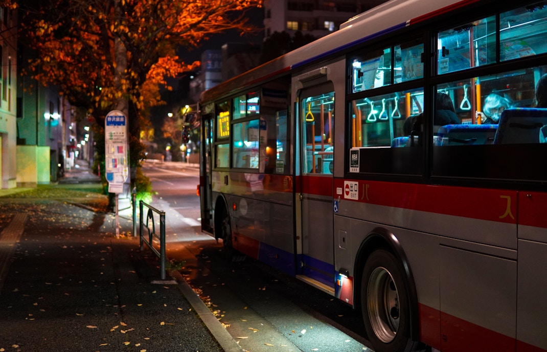 Night bus parked on a city street with illuminated interior and autumn trees.
