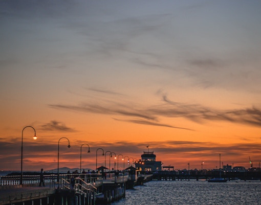 St. Kilda pier at sunset with silhouetted lampposts and calm water.