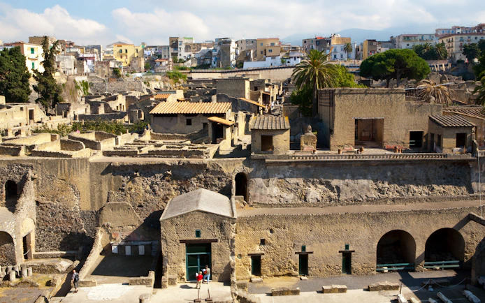 Herculaneum ruins with ancient stone buildings and modern city in the background.