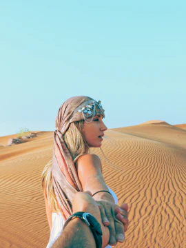 Person leading through Dubai desert dunes, holding hands, with clear sky.