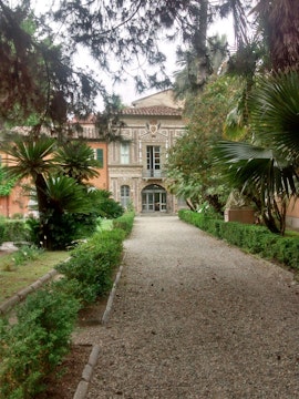 Pathway leading to historic building at Orto Botanico, Palermo, surrounded by lush greenery.