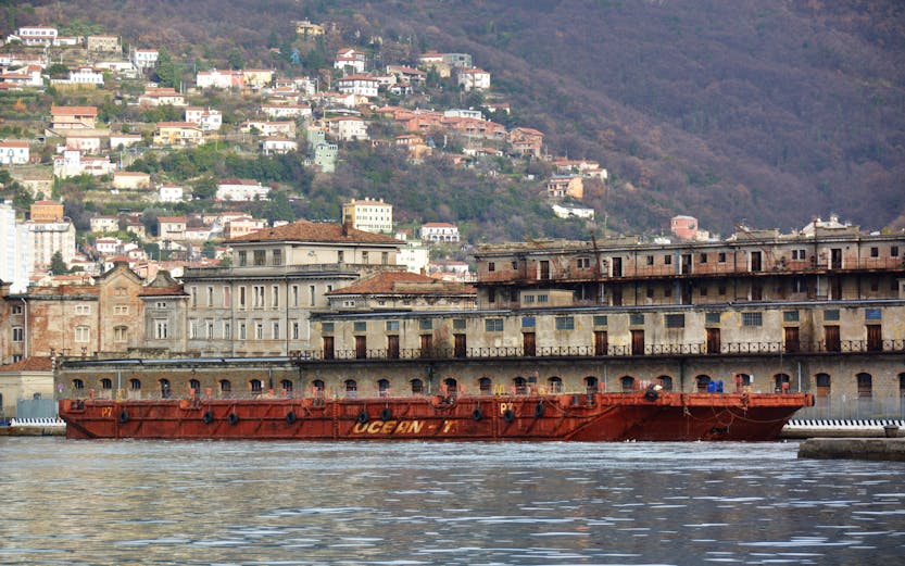 Old port building and ship in Trieste, Italy with hillside houses in the background.