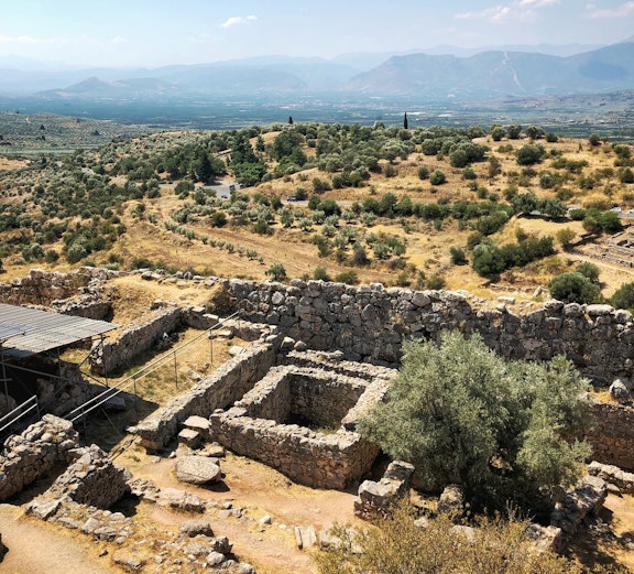 Ancient ruins and olive trees in Delphi, Greece, with mountains in the background.