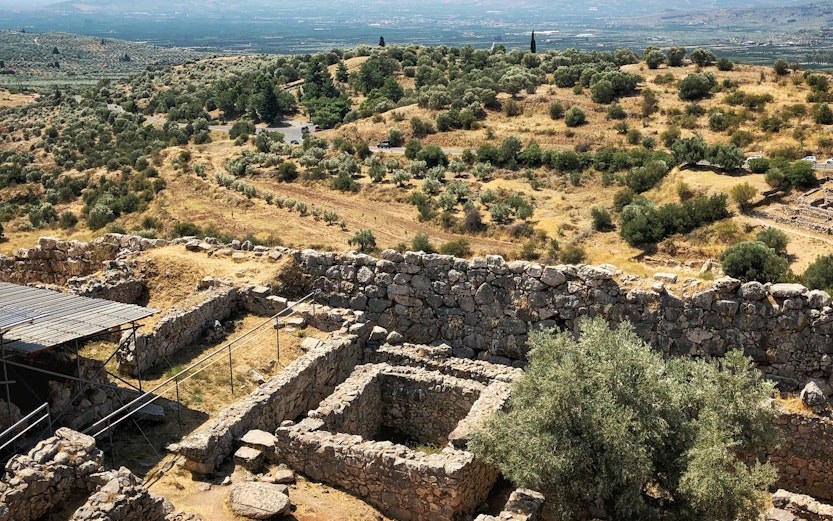 Ancient ruins and olive trees in Delphi, Greece, with mountains in the background.