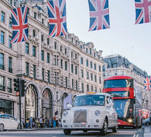 London street with Union Jack flags, classic taxi, and double-decker bus near Oxford & Cambridge tour.