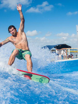 Surfer riding waves on FlowRider with palm trees and spectators in the background.