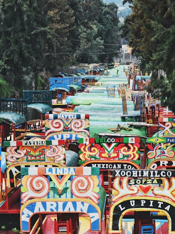 Colorful trajineras on the canal in Xochimilco, Mexico City, surrounded by lush trees.