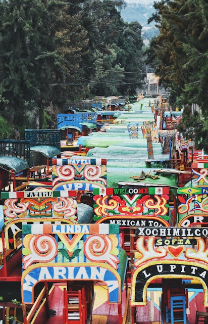 Colorful trajineras on the canal in Xochimilco, Mexico City, surrounded by lush trees.