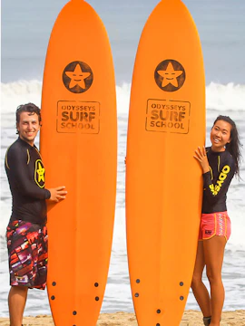 Surfers holding Odysseys Surf School boards on a beach.