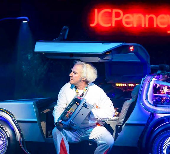 Man in white suit with futuristic car on stage, Olivier Award Winners.
