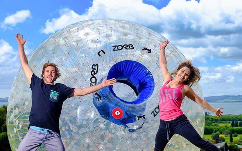 People jumping in front of a Zorb ball on a grassy hill in Rotorua, New Zealand.