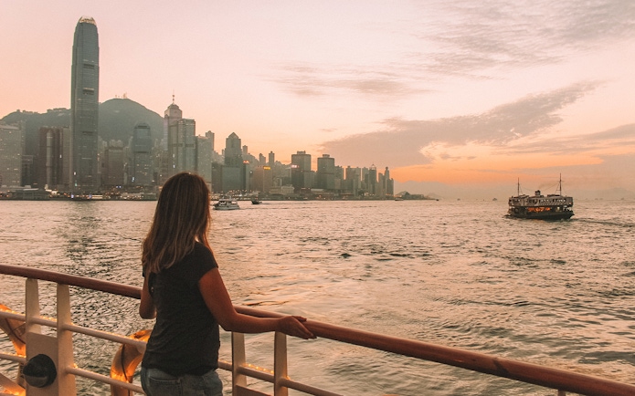 Hong Kong skyline at sunset viewed from a ferry, with a woman looking out over the water.