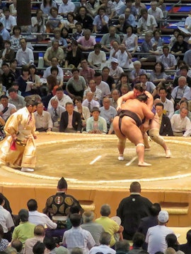 Sumo wrestlers competing in a Fukuoka tournament with a referee and audience watching.