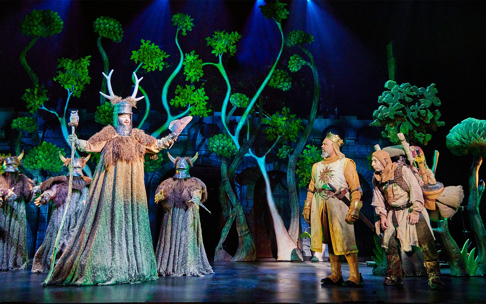 Stage scene from Spamalot with actors in medieval costumes and forest backdrop.