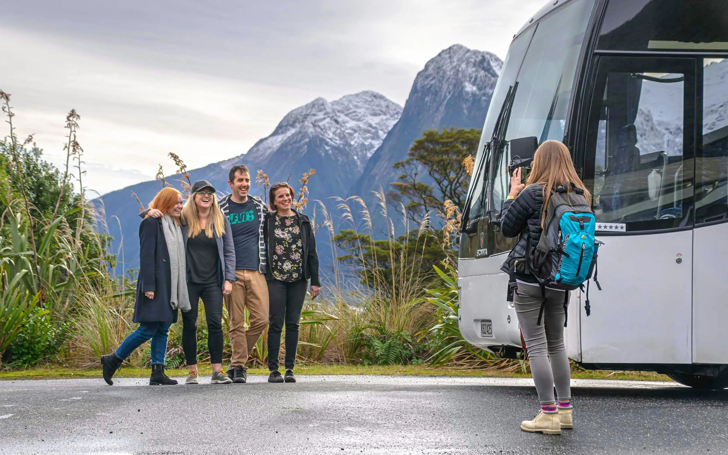Tourists posing for a photo with a bus and mountains in Milford Sound, New Zealand.