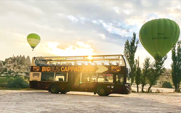 Open-top bus with Cappadocia signage near hot air balloons and rock formations.