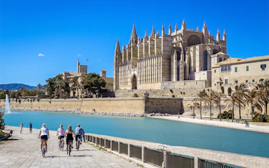 Cyclists near the exterior of Palma Cathedral, Mallorca, with a view of the waterfront.