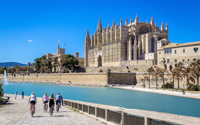 Cyclists near the exterior of Palma Cathedral, Mallorca, with a view of the waterfront.