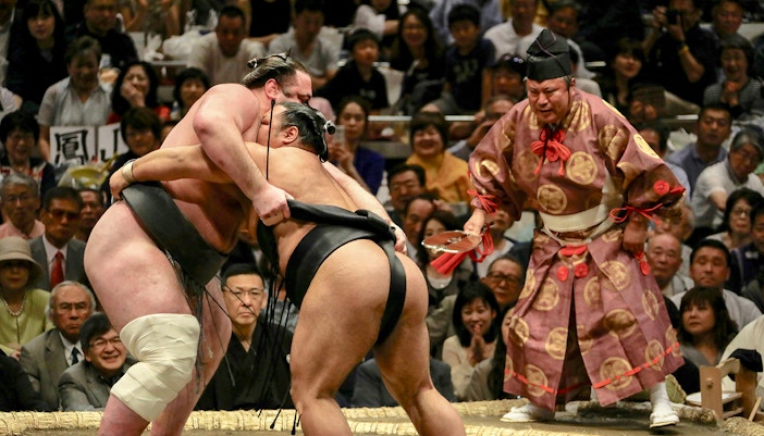 Sumo wrestlers competing at the Tokyo Grand Sumo Tournament with a referee observing.