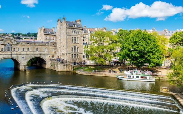 Historic Bath city street with tour group near Pulteney Bridge, live music performance nearby.
