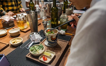 Person preparing Vietnamese coffee-themed cocktail with herbs and spices on a wooden table.