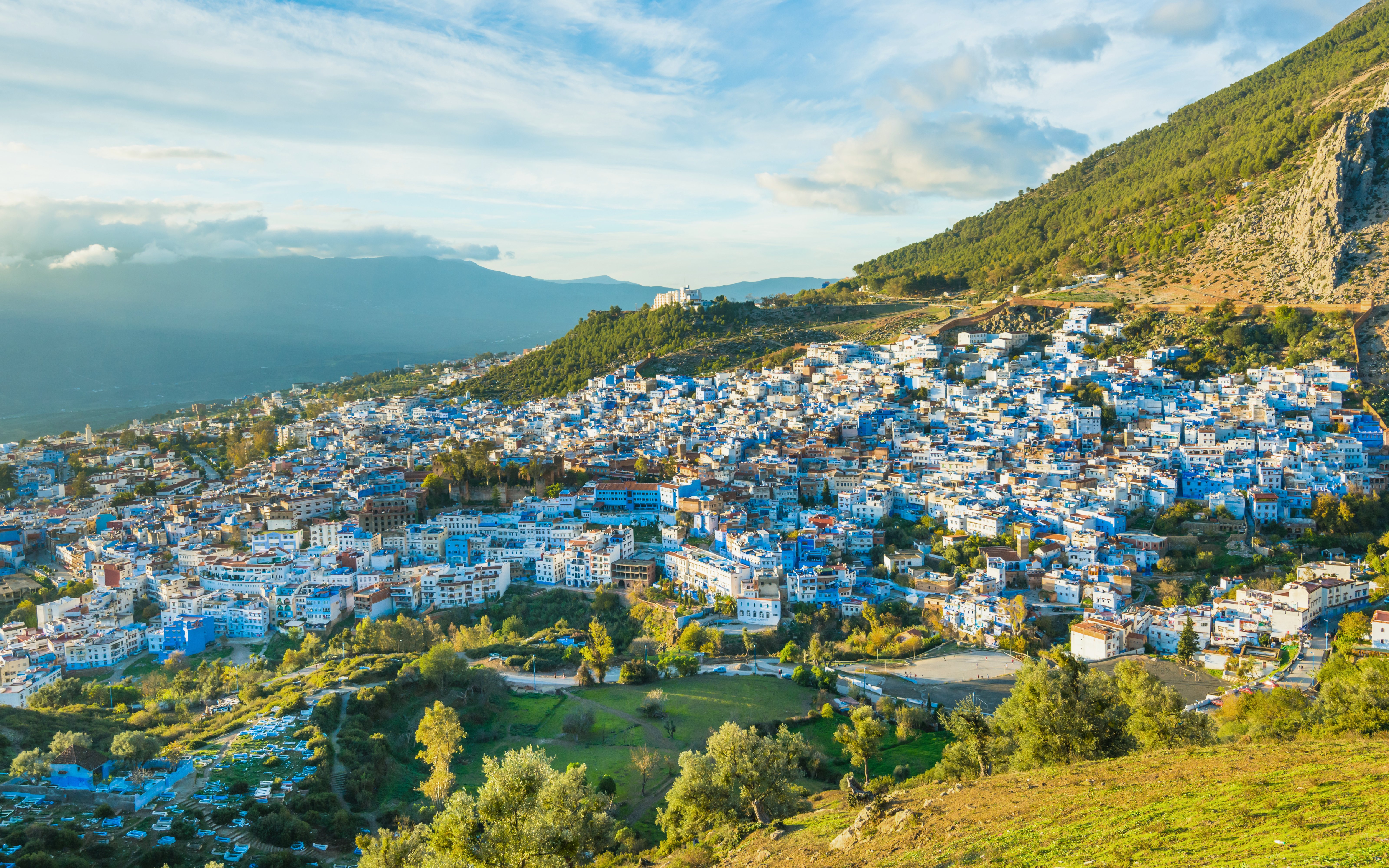 Chefchaouen cityscape with blue buildings, surrounded by green hills and a cloudy sky in Morocco.
