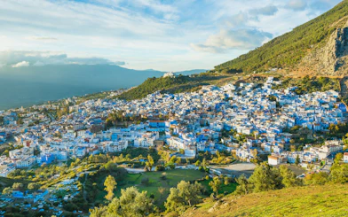 Chefchaouen cityscape with blue buildings, surrounded by green hills and a cloudy sky in Morocco.