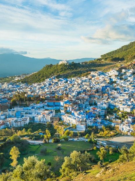 Chefchaouen cityscape with blue buildings, surrounded by green hills and a cloudy sky in Morocco.