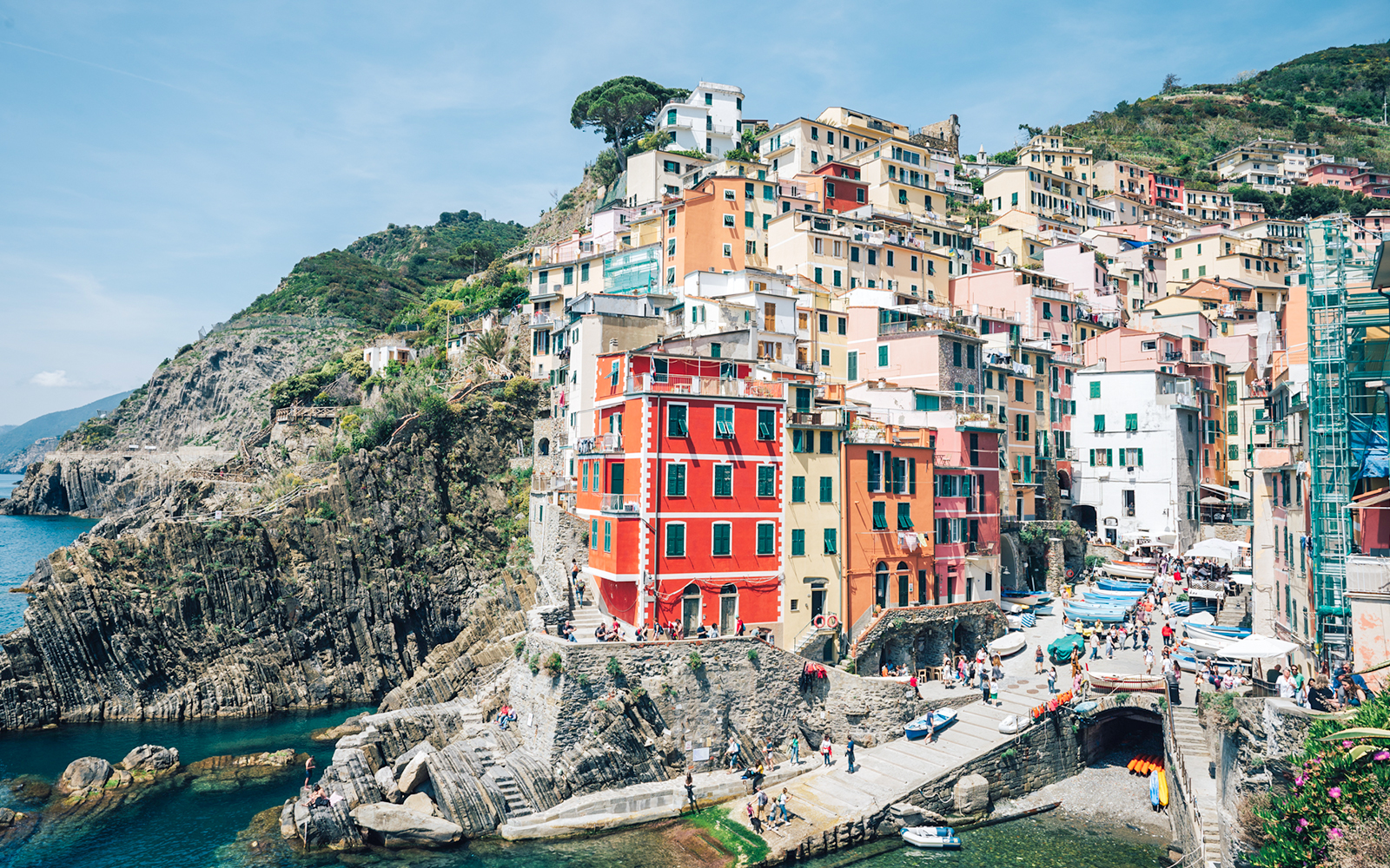 Colorful buildings on cliffs in Riomaggiore, Cinque Terre, Italy, with tourists and boats.