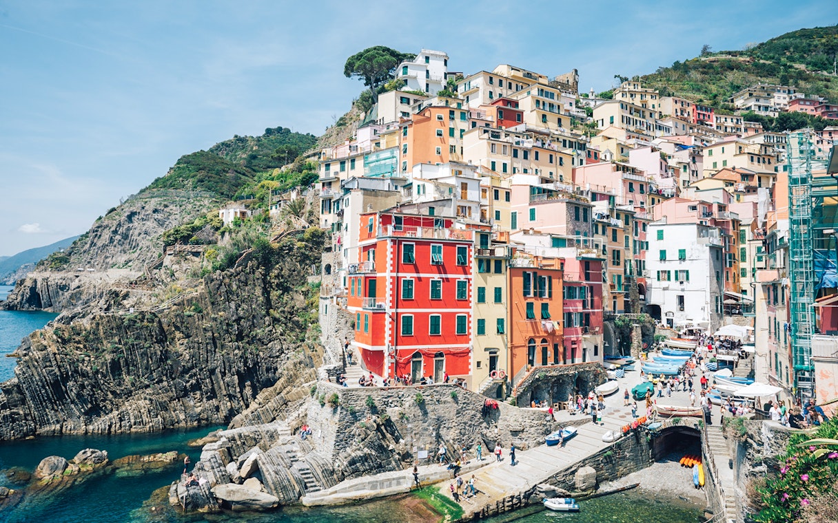 Colorful buildings on cliffs in Riomaggiore, Cinque Terre, Italy, with tourists and boats.