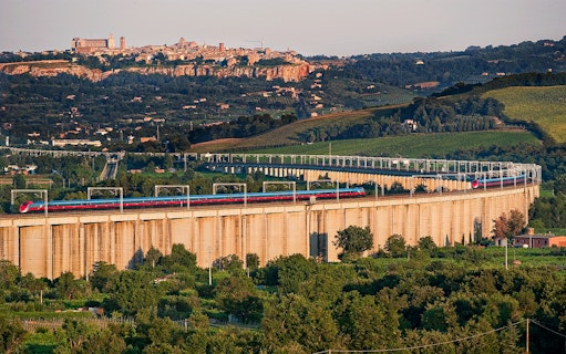 High-speed train crossing a viaduct with Italian countryside and hilltop town in the background.