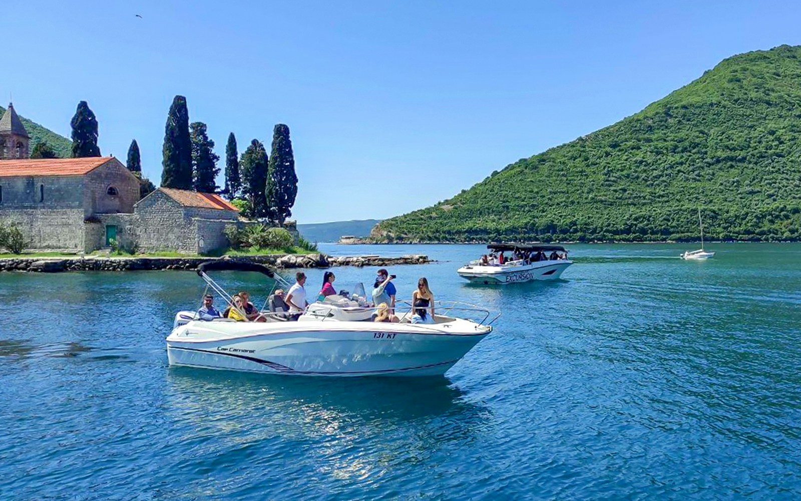 Guests on speedboats near Blue Caves, Montenegro, with a historic stone building and lush hills in the background.