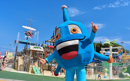 Mascot character at Illa Fantasia Water Park, Barcelona, with water slides and fountains in the background.
