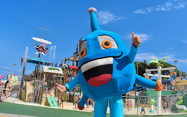 Mascot character at Illa Fantasia Water Park, Barcelona, with water slides and fountains in the background.