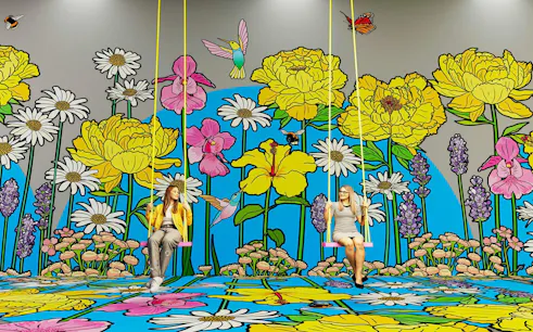 Two people on swings in front of a vibrant floral mural at Bloomyard by Graphic Rewilding.