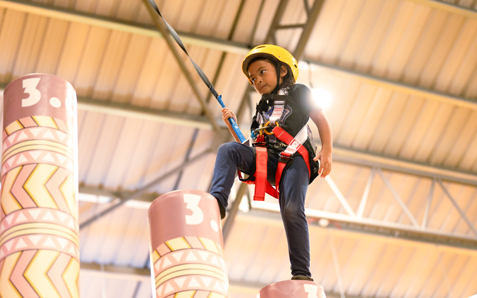 Child balancing on a pole in an indoor activity center, wearing a safety harness and helmet.