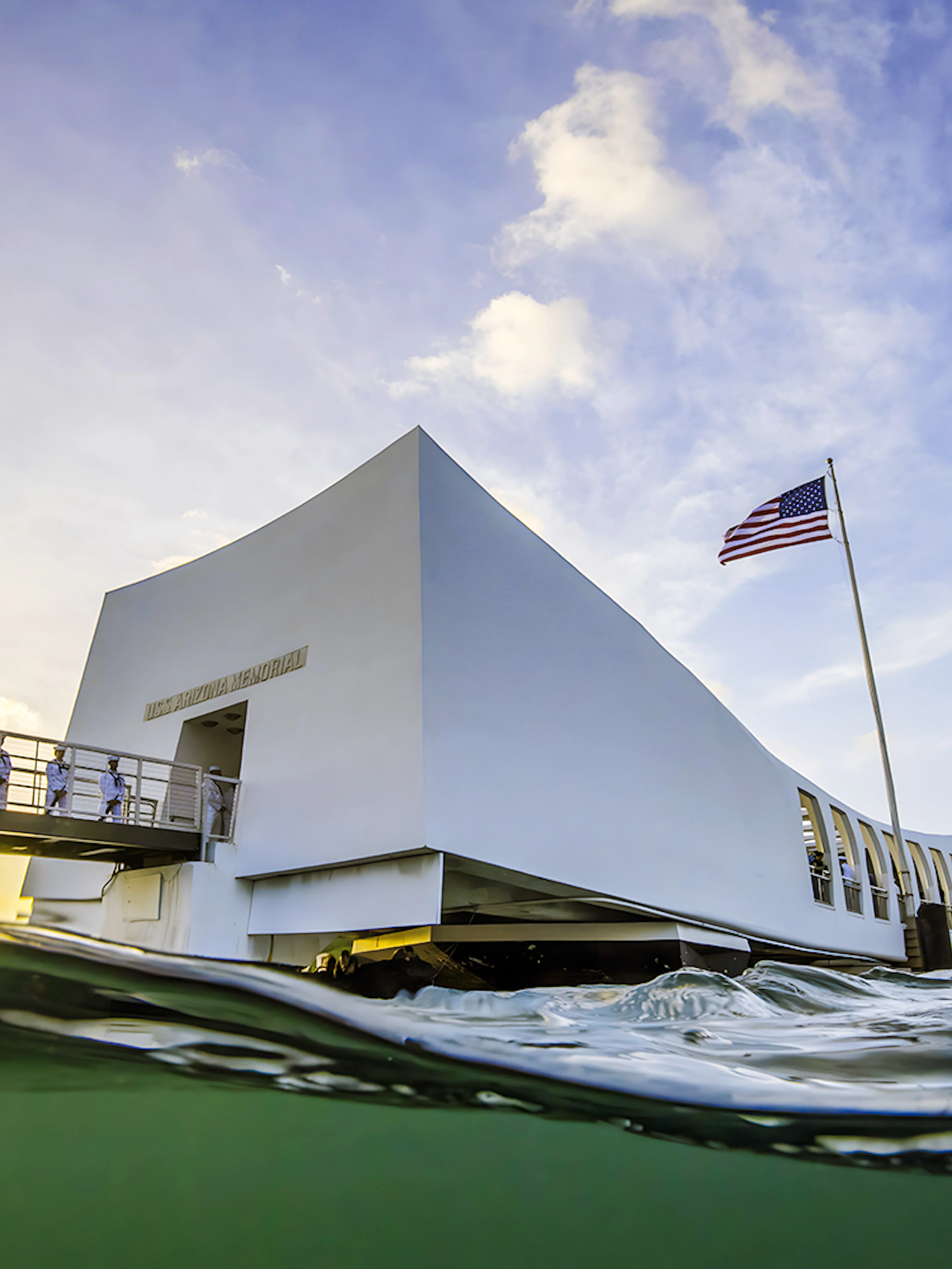 USS Arizona Memorial with American flag and visitors on the bridge in Pearl Harbor, Hawaii.