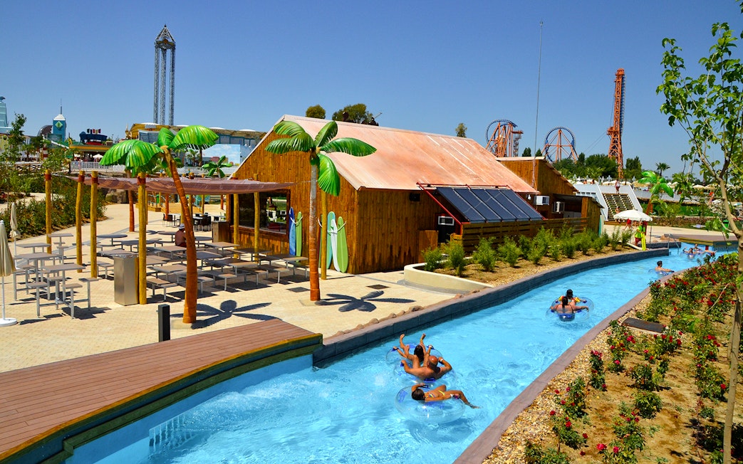 Lazy river with people on tubes at Parque Warner Beach, Madrid, with roller coasters in the background.