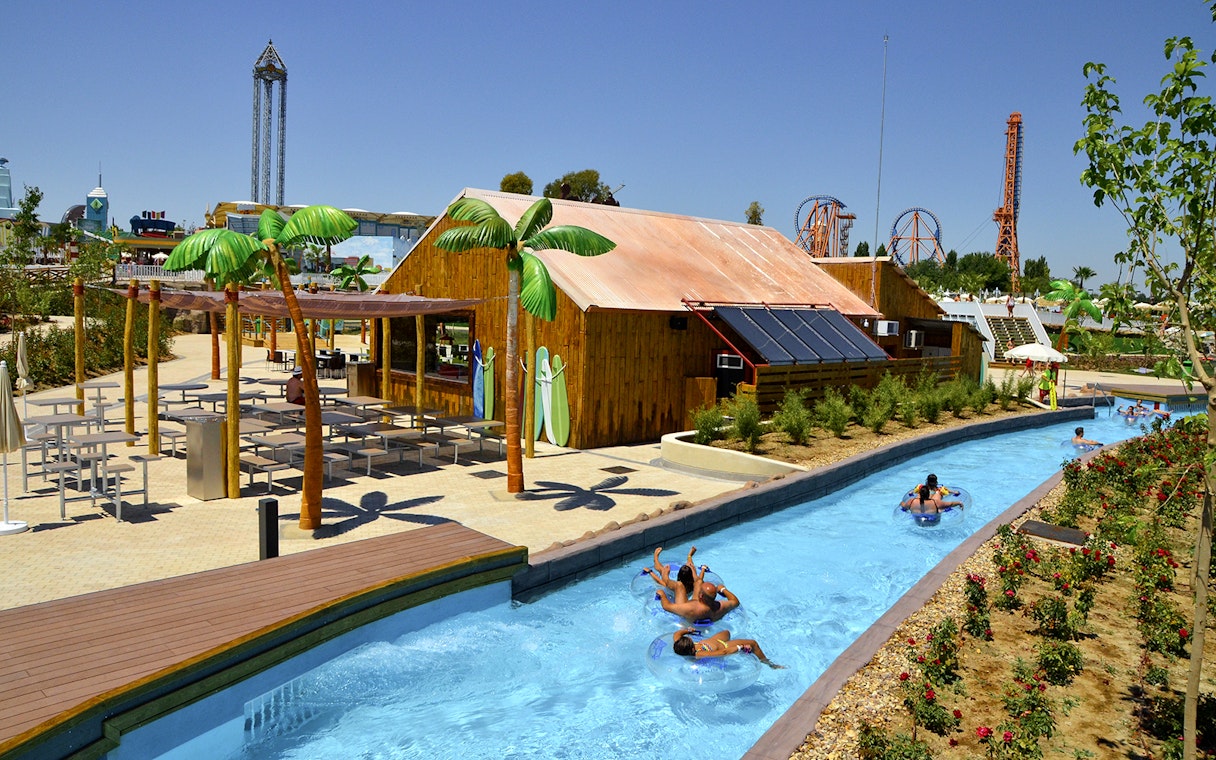 Lazy river with people on tubes at Parque Warner Beach, Madrid, with roller coasters in the background.
